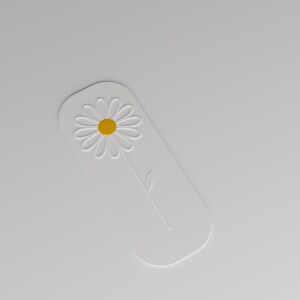 May include: A white rectangular bookmark with rounded corners featuring an embossed daisy flower design. The flower has a bright yellow centre and white petals, with a single embossed stem and leaf extending downwards on the white surface.
