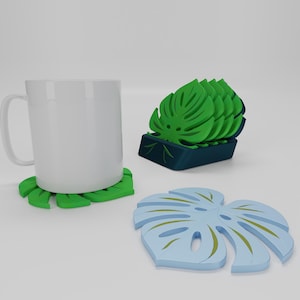 May include: A white mug sits on a green leaf-shaped coaster. Additional monstera leaf-shaped coasters are stacked in a dark blue holder. A single light blue coaster with gold accents is also visible.
