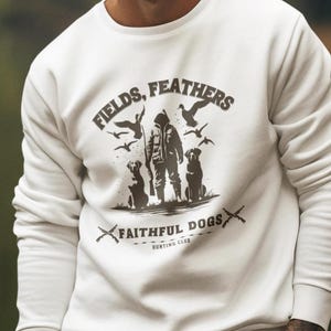 May include: White sweatshirt with the text "FIELDS, FEATHERS" and "FAITHFUL DOGS HUNTING CLUB" in brown. The graphic features a hunter, dogs, and birds. The person is wearing khaki cargo pants.