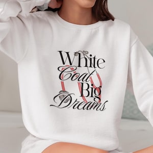 Può includere: Felpa bianca con la scritta "White Coat Big Dreams" in corsivo nero e una grafica di stetoscopio rosso. La felpa è bianca e ha le maniche lunghe. La modella indossa pantaloncini e calzini bianchi.