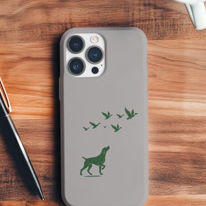 May include: A light grey phone case with a green silhouette of a dog and birds. The case is on a wooden surface, with a pen and a cup of coffee visible.