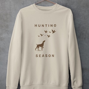 以下が含まれることがあります： ベージュ色のスウェットシャツ。茶色の文字で「HUNTING SEASON」と書かれています。犬の茶色のシルエットが、飛んでいる鳥を見上げており、「HUNTING」の下に配置されています。
