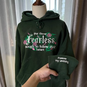 Puede incluir: Sudadera con capucha verde oscuro con el texto bordado "For those Fearless enough to follow their heart" en blanco y rosa. El puño de la manga tiene el texto "Forever my undoing". La sudadera tiene una capucha con cordón.
