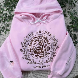 May include: A light pink hoodie with a drawstring hood. The front features a brown embroidered design with the text "THE ARCHER" and "++THE FOX++". The left sleeve has a small peach embroidery.