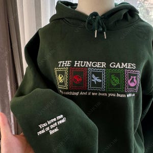 Puede incluir: Sudadera con capucha verde oscuro con el texto "THE HUNGER GAMES" y diseños de sellos bordados coloridos. La manga presenta el texto "You love me real or not real? Real".