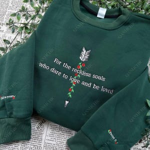 Puede incluir: Sudadera verde oscuro con texto bordado en blanco: "For the reckless souls who dare to love and be loved." El diseño incluye una flecha blanca y detalles florales rojos. La manga tiene el texto "Prove it."