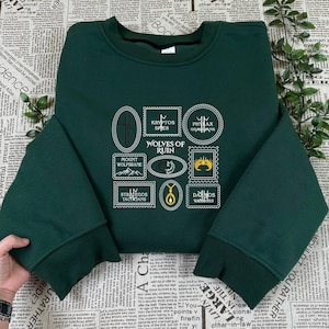 Op de afbeelding: Donkergroene sweatshirt met een ronde hals en lange mouwen. De voorkant heeft een ontwerp van verschillende stempelachtige afbeeldingen met tekst, waaronder "Kryptos Spies" en "Wolves of Ruin".