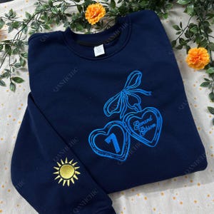 Puede incluir: Sudadera azul marino con diseño de corazón bordado en azul. Un corazón tiene el número 1, el otro el nombre "Gerard Gibson". Un sol amarillo está bordado en la manga. La marca "QANHCHIC" está impresa en la sudadera.