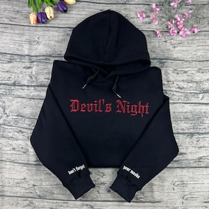 May include: Black hoodie with the text "Devil's Night" in red gothic lettering across the chest. The sleeves have the phrases "don't forget" and "your masks" in white. The hoodie has a drawstring and hood.