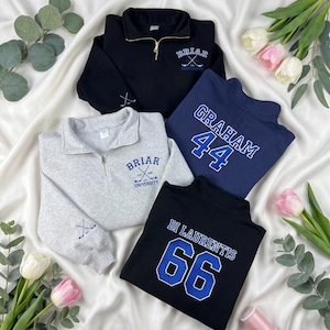 Op de afbeelding: Vier sweatshirts met een kwartrits in zwart, marineblauw en grijs. De sweatshirts hebben geborduurde tekst en afbeeldingen, waaronder "Briar University" en spelersnamen met nummers. De sweatshirts worden gepresenteerd op een witte stof met bloemen.