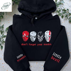 May include: Black hooded sweatshirt with embroidered mask designs in red and white. The text "don't forget your masks" is below the masks. The sleeves have the phrases "little trouble" and "little devil".