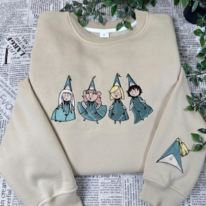 Könnte beinhalten: Ein beigefarbenes Sweatshirt mit Rundhalsausschnitt und einer Stickerei von vier Zeichentrickfiguren, die spitze, blaugrüne Hüte und passende Umhänge tragen. Ein einzelner spitzer, blaugrüner Hut mit gelber Quaste ist auf dem linken Ärmel gestickt.