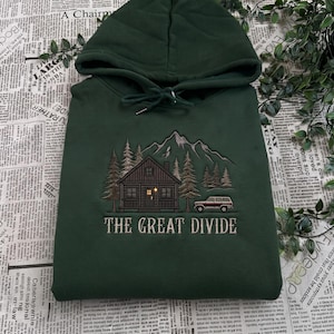 Könnte beinhalten: Ein dunkelgrüner Kapuzenpullover mit einer Stickerei, die eine Hütte im Wald mit Bergen im Hintergrund und einem geparkten Fahrzeug zeigt. Der Text THE GREAT DIVIDE ist in einer hellen Farbe unter der Hüttenszene eingestickt.