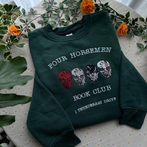 May include: Dark green sweatshirt with the text "FOUR HORSEMEN BOOK CLUB 1 THUNDERBAY DRIVE". The sweatshirt features four embroidered skull designs in red, gray, black, and white. The sweatshirt is made of a soft material.
