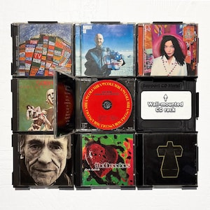 Puede incluir: Un estante de CD montado en la pared, que muestra varias portadas de álbumes. El estante es negro y contiene nueve estuches de CD. Las portadas incluyen imágenes de músicos, obras de arte y texto. Un estuche está abierto, revelando un CD rojo. El texto dice "Wall-mounted CD rack".