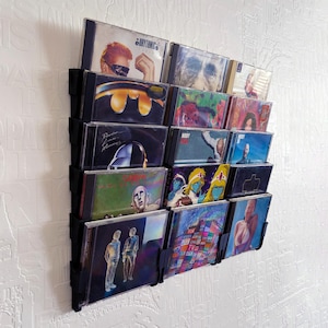 Peut inclure: Support mural noir pour CD rempli de divers CD de musique. Le support est en plastique noir et contient plusieurs CD dans des fentes individuelles. Les CD présentent des pochettes d'album avec des couleurs et des motifs variés.