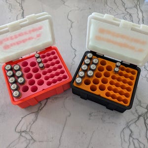 Könnte beinhalten: Zwei offene Batteriestaukästen. Einer ist rot mit rosa Innenraum, der andere schwarz mit orangefarbenem Innenraum. Jede Box enthält mehrere AA-Batterien. Die Deckel sind weiß und leicht durchscheinend.