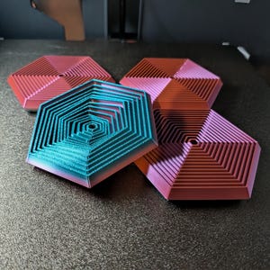 Può includere: Set di sottobicchieri esagonali nei toni del rosa, rosso e turchese. I sottobicchieri presentano un design a strati e a gradini. Il sottobicchiere turchese ha un effetto sfumato, che passa dal turchese al rosa.