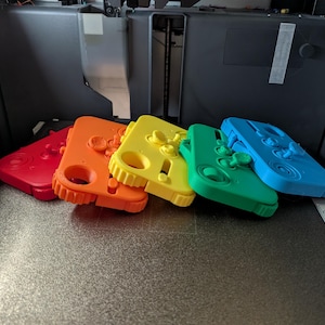 May include: A collection of five brightly colored, 3D-printed game controller cases in red, orange, yellow, green, and blue. Each case features a unique button and joystick design, and is angled on a gray surface.