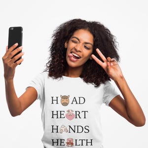 May include: White t-shirt with the words "HEAD HEART HANDS HEALTH" and animal illustrations. A person is holding a black smartphone and making a peace sign with their fingers.