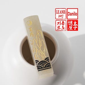 Może przedstawiać: Prostokątny, kremowy kamienny stempel ze złotymi wzorami gór i fal. Stempel ma chińskie znaki i symbol księżyca. Stempel umieszczony jest w białym ceramicznym pojemniku. Dwa czerwone stemple z tekstem znajdują się w prawym górnym rogu.