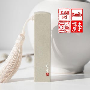 Puede incluir: Un sello rectangular de piedra beige con una borla blanca. El sello tiene texto coreano y un sello cuadrado rojo en la parte inferior. El fondo incluye un jarrón blanco y sellos rojos adicionales con texto.