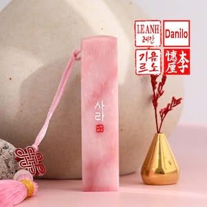 May include: A pink rectangular stamp with Korean text, a pink cord, and a decorative tassel. The stamp has a marbled appearance. The background includes a gold vase with red stems and a beige ceramic object. The image also includes two red stamps with Korean text.