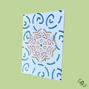 May include: A white card with a central mandala design in gold and red glitter, surrounded by teal swirls and lines. The card has a slight angle, showcasing the design's details. The bottom right corner features a small logo.