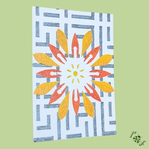 May include: A white card with a geometric pattern in gray and a central floral design. The flower features orange and yellow petals surrounding a yellow sun-like center. The card is angled, showing its design.