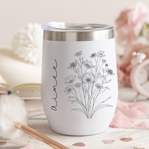 Op de afbeelding: Een witte roestvrijstalen beker met een doorzichtig deksel, voorzien van een zwarte lijntekening van een boeket wilde bloemen en de naam Aimee verticaal geschreven in een cursief lettertype op de zijkant van de beker.