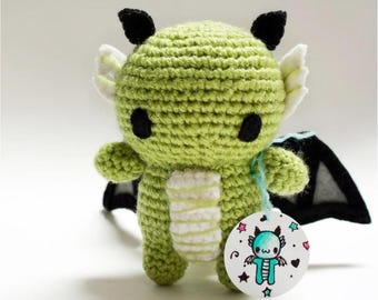 May include: A handmade green crochet dragon with black eyes, ears, and wings. The dragon has white accents and a white and yellow belly. A round tag with a dragon illustration and stars is attached.