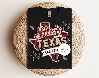 May include: A black t-shirt featuring a graphic of the state of Texas in red glitter. The text reads She's from TEXAS I CAN TELL, with a small white circular badge that says CUSTOM MASCOT. The design includes small star accents in white and gold.