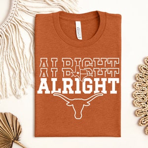 May include: A burnt orange t-shirt with the text "AT DIGHT AT DIGHT ALRIGHT" in white. The text is stacked vertically with a white outline of the state of Texas and a longhorn steer below the text.