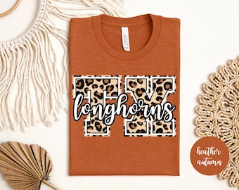 Op de afbeelding: Een oranje T-shirt met een witte "TX Longhorns" afbeelding. De letters zijn gevuld met een luipaardprint.