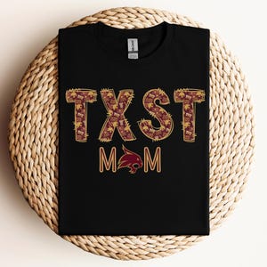 May include: Black t-shirt with the text "TXST MOM" in maroon and gold lettering. The letters are filled with a pattern. Below the text is a maroon bobcat logo.