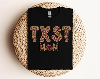 May include: Black t-shirt with the text "TXST MOM" in maroon and gold lettering. The letters are filled with a pattern. Below the text is a maroon bobcat logo.
