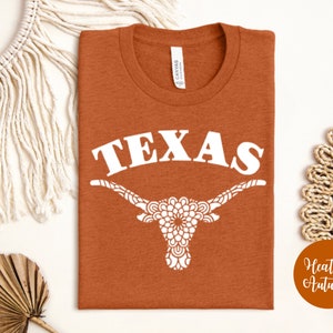 May include: A rust-colored t-shirt with a white graphic of a Texas longhorn with a mandala design and the word "TEXAS" above it. The t-shirt is on a white background.