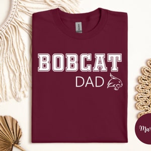 May include: Maroon t-shirt with white text that reads "Bobcat Dad" and a white bobcat mascot graphic.