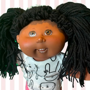 Może przedstawiać: Lalka Cabbage Patch Kids z ciemną skórą, brązowymi oczami i czarnymi włosami z przędzy w kucykach. Lalka ma na sobie białe body z czarnym nadrukiem królika i różowe spodnie. Widoczne jest logo Cabbage Patch Kids.