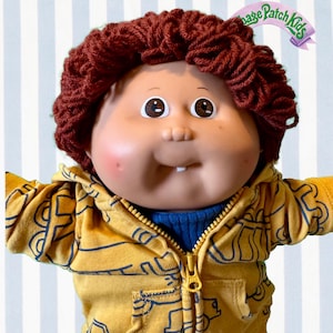 Puede incluir: Muñeca Cabbage Patch Kids con cabello de hilo marrón, que lleva una sudadera con capucha amarilla con cremallera con un diseño azul y un suéter azul debajo. La muñeca tiene mejillas rosadas y un diente pequeño. El logotipo de Cabbage Patch Kids es visible.