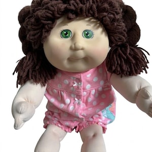 May include: A doll with a soft fabric body, a round face, and green eyes. The doll has thick, dark brown yarn hair styled in pigtails. It is wearing a pink romper featuring a white polka dot pattern and small white bows on the front bodice.