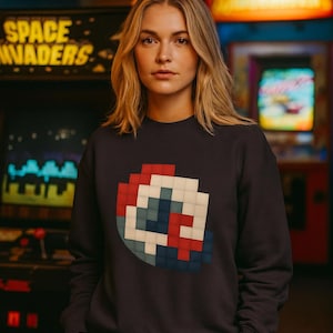 May include: A dark navy blue sweatshirt featuring a pixelated graphic design in red, white, and blue. The sweatshirt is worn in an arcade setting, with the words "SPACE INVADERS" visible in the background.