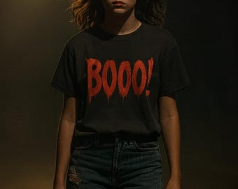 May include: A person wearing a dark gray t-shirt with the word "BOOO!" in dripping red letters. The person is also wearing blue jeans with distressed details. The background is dark with a single overhead light.
