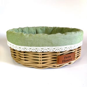 Puede incluir: Una cesta redonda de mimbre trenzado con un forro de tela verde salvia y un borde de encaje blanco en la parte superior. En el lateral lleva una etiqueta rectangular marrón de aspecto cuero con el texto Fern Flower en letras negras.