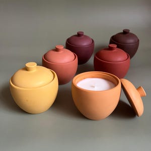 Puede incluir: Conjunto de recipientes de cerámica con forma de bellota en varios tonos tierra, incluyendo amarillo, naranja, rojo y marrón. Uno de los recipientes está abierto, revelando una vela blanca. Las tapas son redondas con un pequeño pomo. Fondo verde apagado.