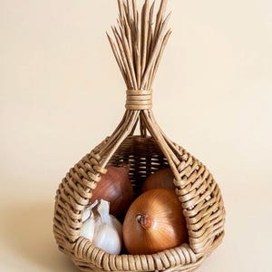 May include: A woven wicker basket with a teardrop shape, filled with onions and garlic. The basket has a handle made of bundled twigs. The onions are brown and red, and the garlic is white. The basket is a natural beige color.