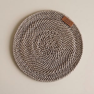 May include: A circular placemat with a woven spiral design, in a light brown hue. A small, rectangular leather tag with the words "Fern Flower" is attached. The placemat is made from natural materials, suitable for dining.