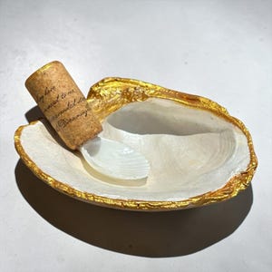 May include: A white oyster shell dish with a gold-painted rim. Inside the shell rests a cylindrical cork stopper featuring printed black text, which is attached to a small, white, ridged seashell scoop that sits within the larger shell bowl.
