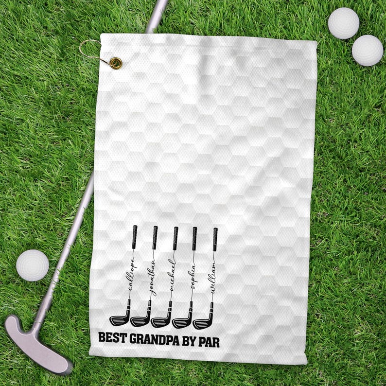 May include: White golf towel with a honeycomb pattern and the text "BEST GRANDPA BY PAR". The towel features golf club illustrations with names: Callie, Jonathan, Michael, Sophia, and William. Golf balls and a putter are also visible.