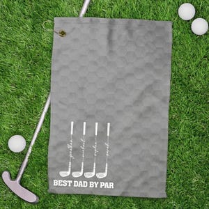 May include: A grey golf towel with a honeycomb pattern, golf clubs, and the text "BEST DAD BY PAR." The names Jonathan, Michael, Sophia, and Sarah are printed on the golf clubs. Two golf balls and a putter are on the green grass.